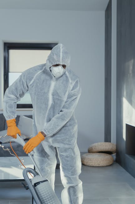 A person dressed in light-colored overalls is performing deep cleaning or surface sanitisation on a carpeted floor using a professional vacuum or extraction machine at an indoor location, possibly a c