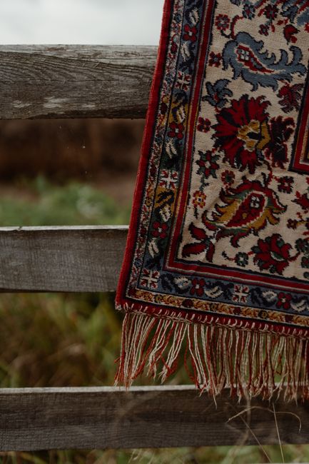 Close-up of a patterned oriental rug with intricate floral and geometric designs in red, blue, yellow, and cream colors hanging over a weathered wooden fence outdoors. The rug's fringed edge is visibl
