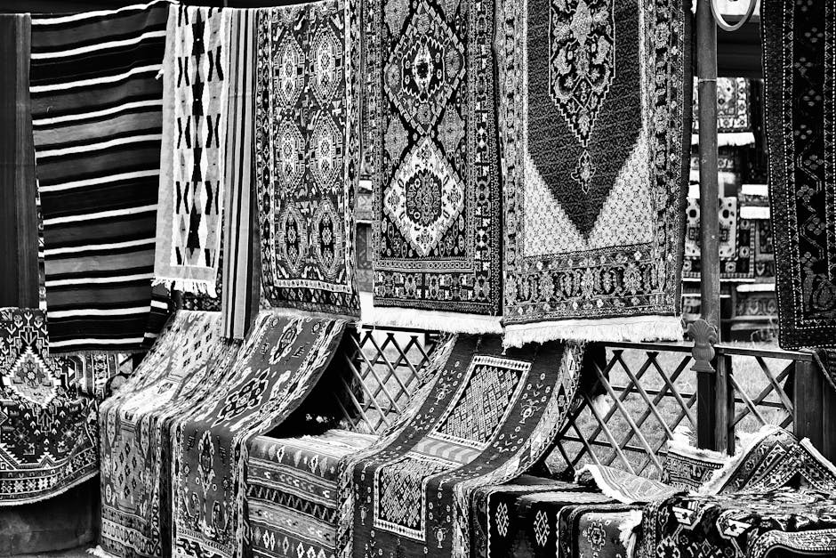 Close-up of a patterned oriental rug with intricate floral and geometric designs in red, blue, yellow, and cream colors hanging over a weathered wooden fence outdoors. The rug's fringed edge is visibl